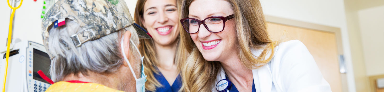 Two women smiling as they talk with an older man who is wearing a baseball cap and a mask on his face